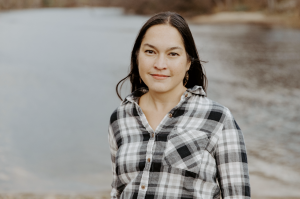 Sachi is smiling at the camera wearing a black and white plaid button down shirt. She is standing in front of a river.